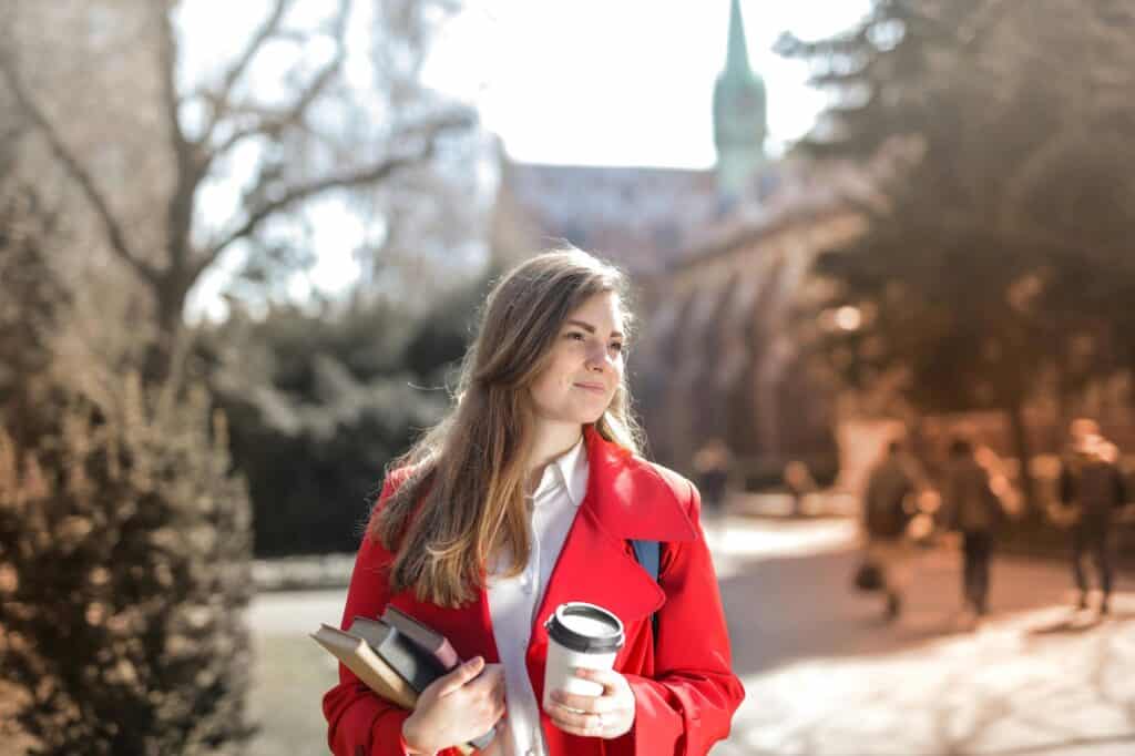 A female student is walking inside the campus with her books and a cup of coffee.