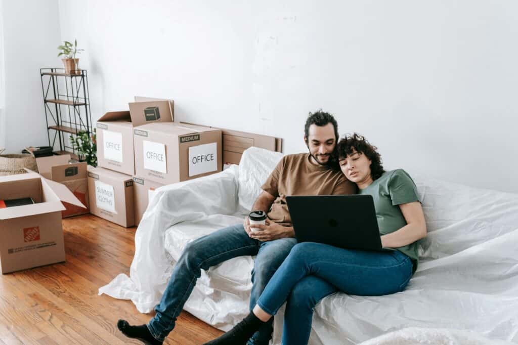 A couple sitting on a wrapped sofa and surrounded by cartons in the concept of affordable housing options when moving to Romford.